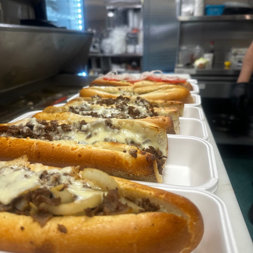 A row of Amore Pizza and Pasta's Philly cheesesteaks with melted cheese and meat in a kitchen.